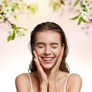 Naturalna kobieta z zamkniętymi oczami i uśmiechem, dotyka policzków dłońmi, nad nią gałązki kwitnącej wiśni. 