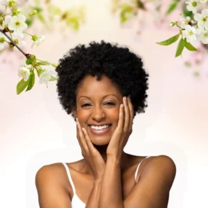 Roześmiana kobieta, dotyka dłońmi twarzy a nad nią kwitnące gałązki.