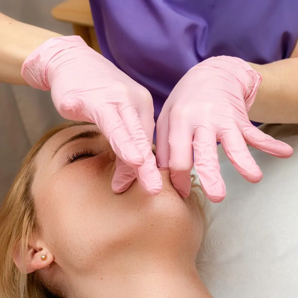 Kobieta, blondynka na masażu transbukalnym (wewnątrzpoliczkowym). Dłonie masażysty, w różowych rękawiczkach stymulują mięśnie okrężne ust. Masażystka ma fioletowy uniform.