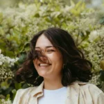 Naturalna, uśmiechnięta dziewczyna, brunetka z rozwianymi włosami do ramion, na tle zieleni.